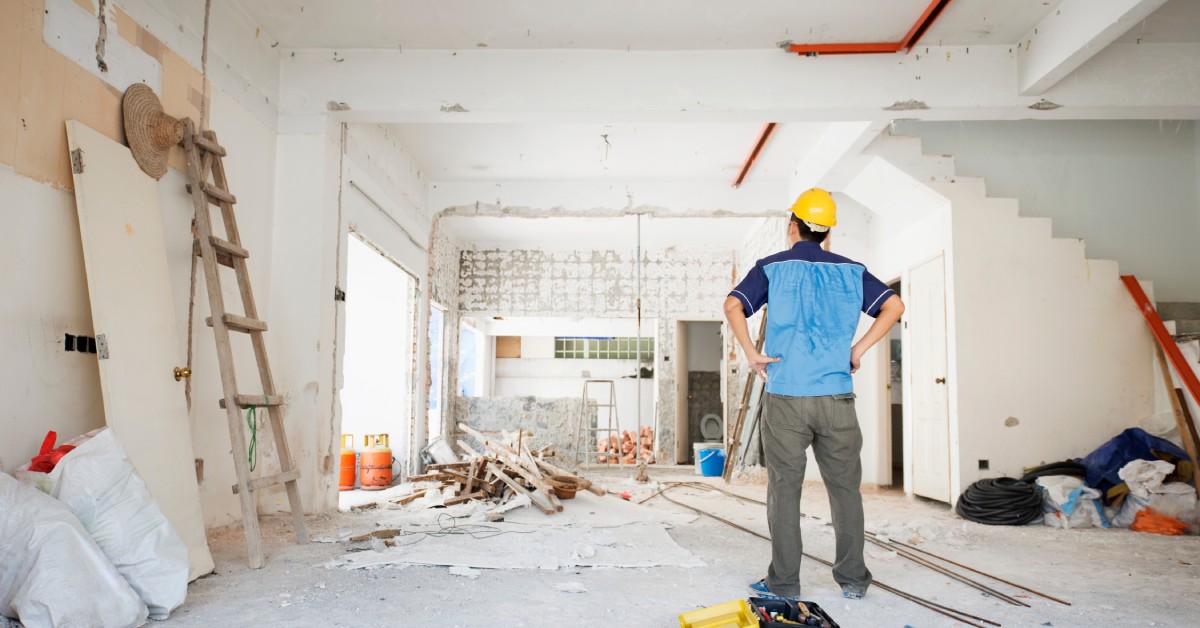 A contractor wearing a blue shirt and yellow hard hat stands in the middle of a house under construction and looks up.