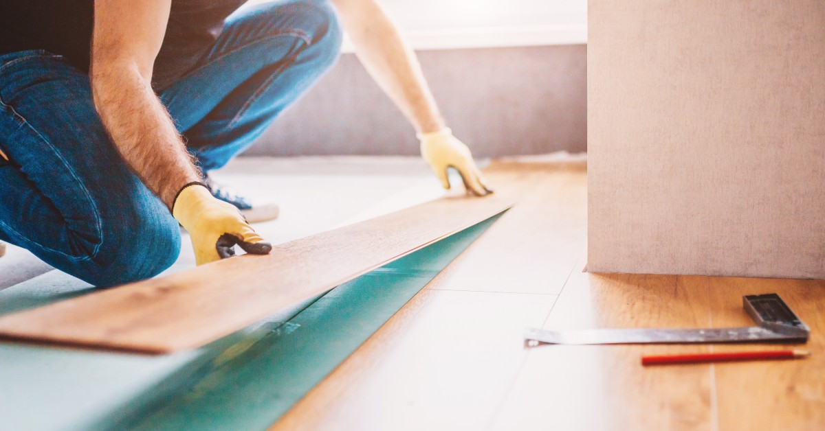 A worker wearing yellow gloves and jeans installs a tan flooring plank. A pencil and a square tool lie on the floor.