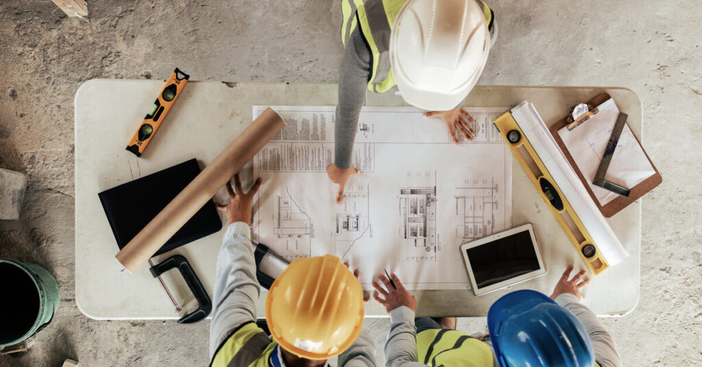 An overhead view of three professional contractors gathered around a table and discussing home design blueprints.