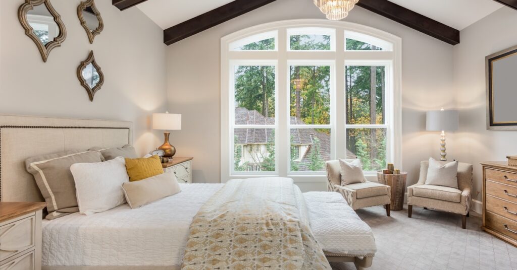 A beautiful bedroom featuring a comfy bed, pillows, chairs, brown wooden beams, mirrors, and a chandelier.