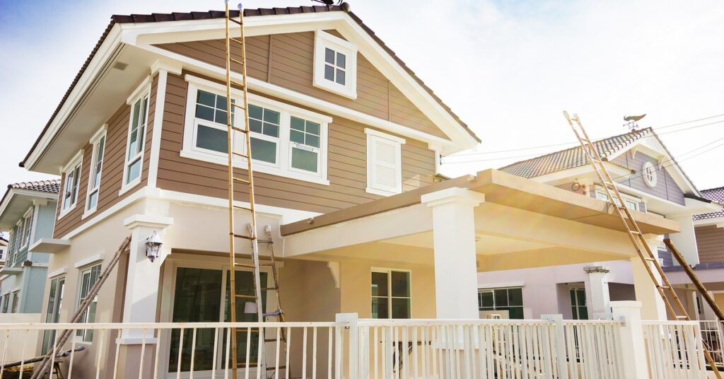 A home's exterior showing signs of a renovation with various ladders outside. The sky in the background has white clouds.