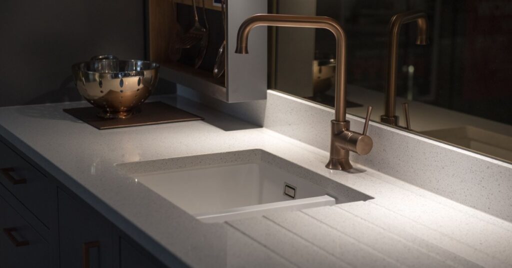 A home's kitchen counter features a single bowl sink with a bronze faucet next to a window. The counter is white.