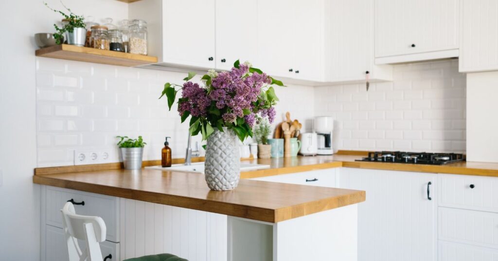 A small home's kitchen features counters and stove spaces. A potted plant with purple flowers sits on the counter.